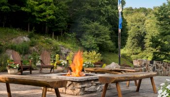 Outdoor patio with a fire pit, wooden chairs, a flagpole, and lush greenery in the background.
