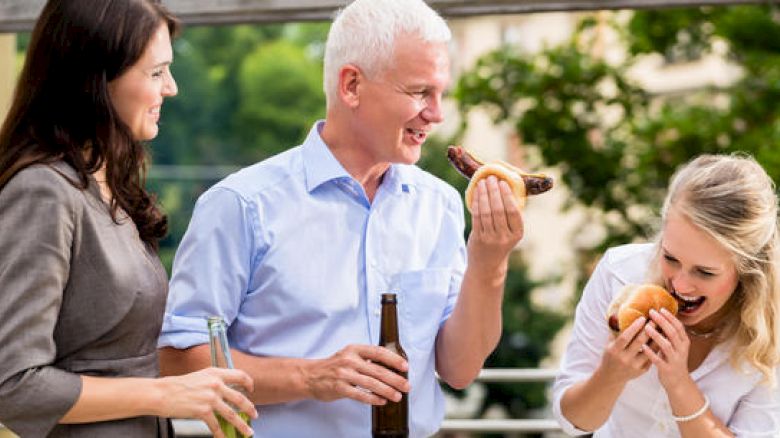 Three people are enjoying a barbecue together, with two holding beers and one eating a hot dog.