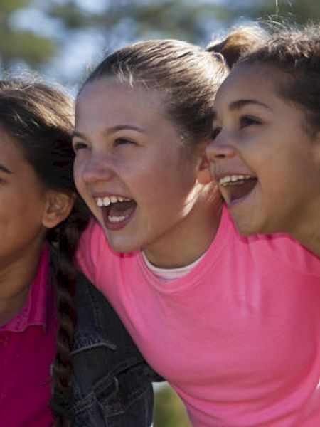 Three girls smiling outdoors, with one embracing the others in a show of friendship.