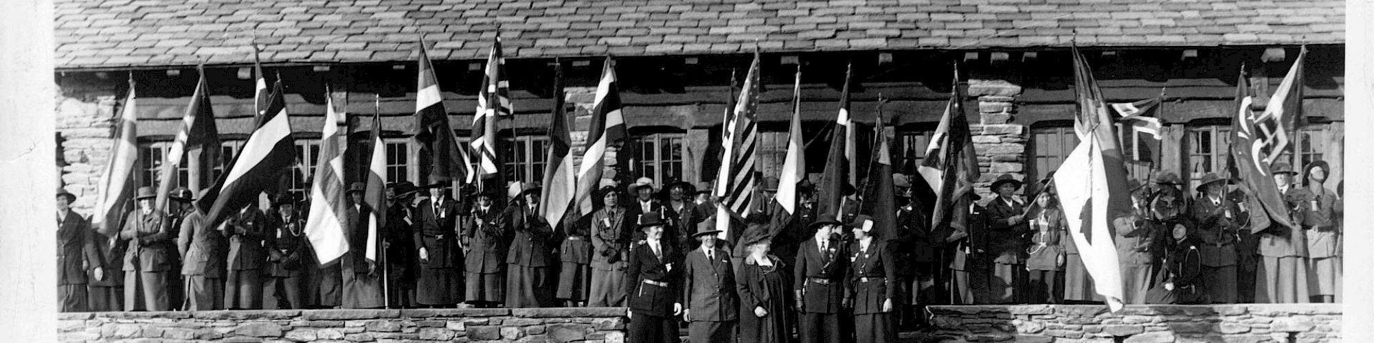 Black and white photo of people in uniforms holding flags in front of a rustic building.