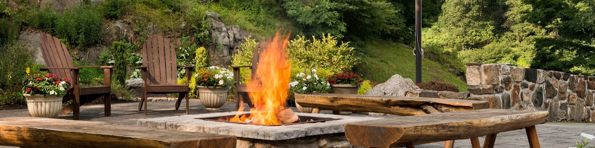 An outdoor fire pit is lit, surrounded by wooden Adirondack chairs, flagpole to the side, all set against a verdant backdrop.