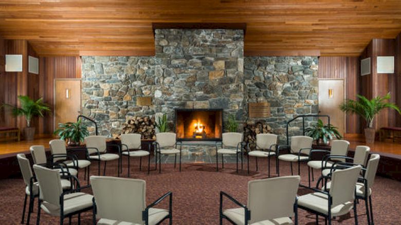 A cozy room with a lit fireplace, stone wall, wood ceiling, chairs arranged in a circle.