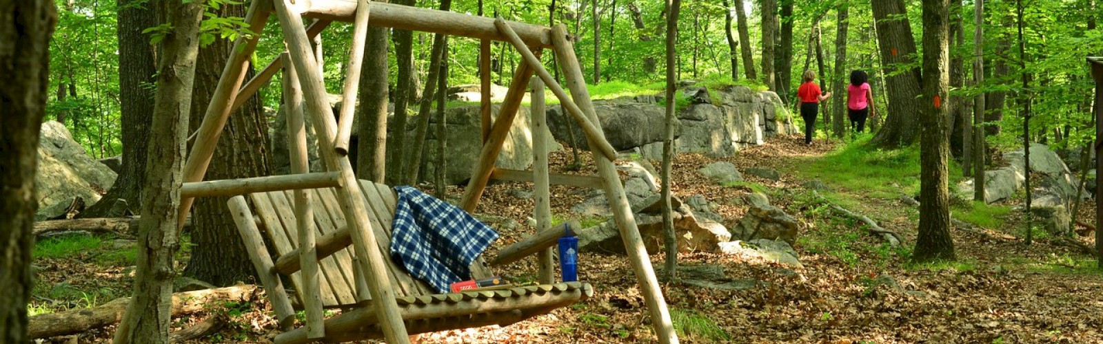 A wooden swing with a blanket in a forest, two people in the distance walking.