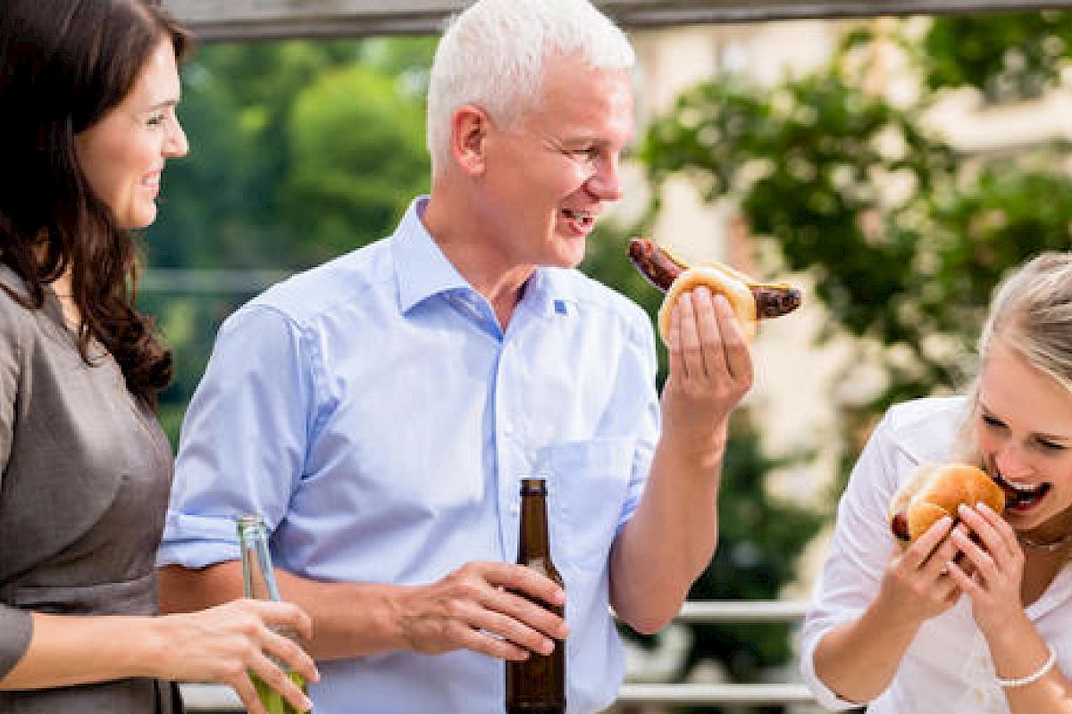 Three people are enjoying a barbecue together, with two holding beers and one eating a hot dog.