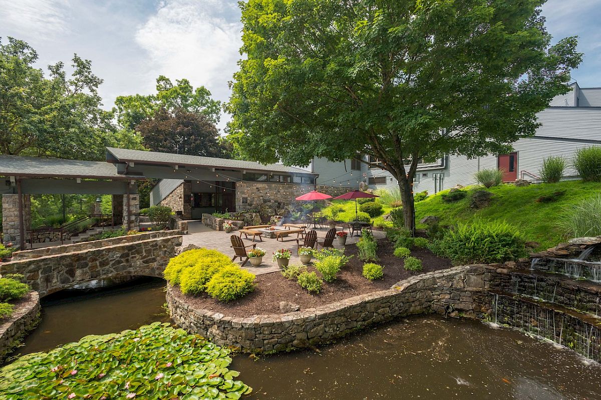 Serene garden with pond, lush trees, a patio with furniture, and a red umbrella.