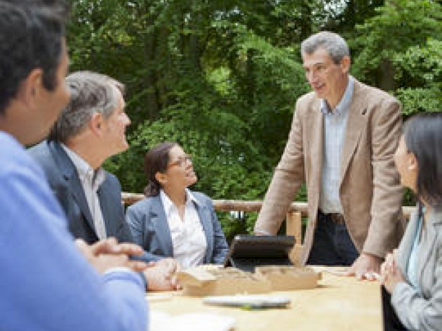 Group of people having a meeting outdoors, some seated, one standing.
