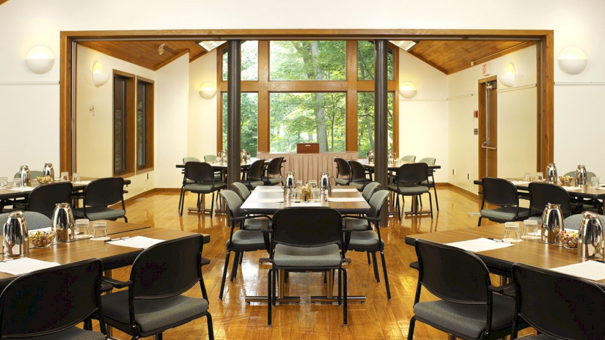A spacious room with wooden floors, set up for a meeting or event. Tables and chairs are arranged symmetrically, with a scenic view outside.