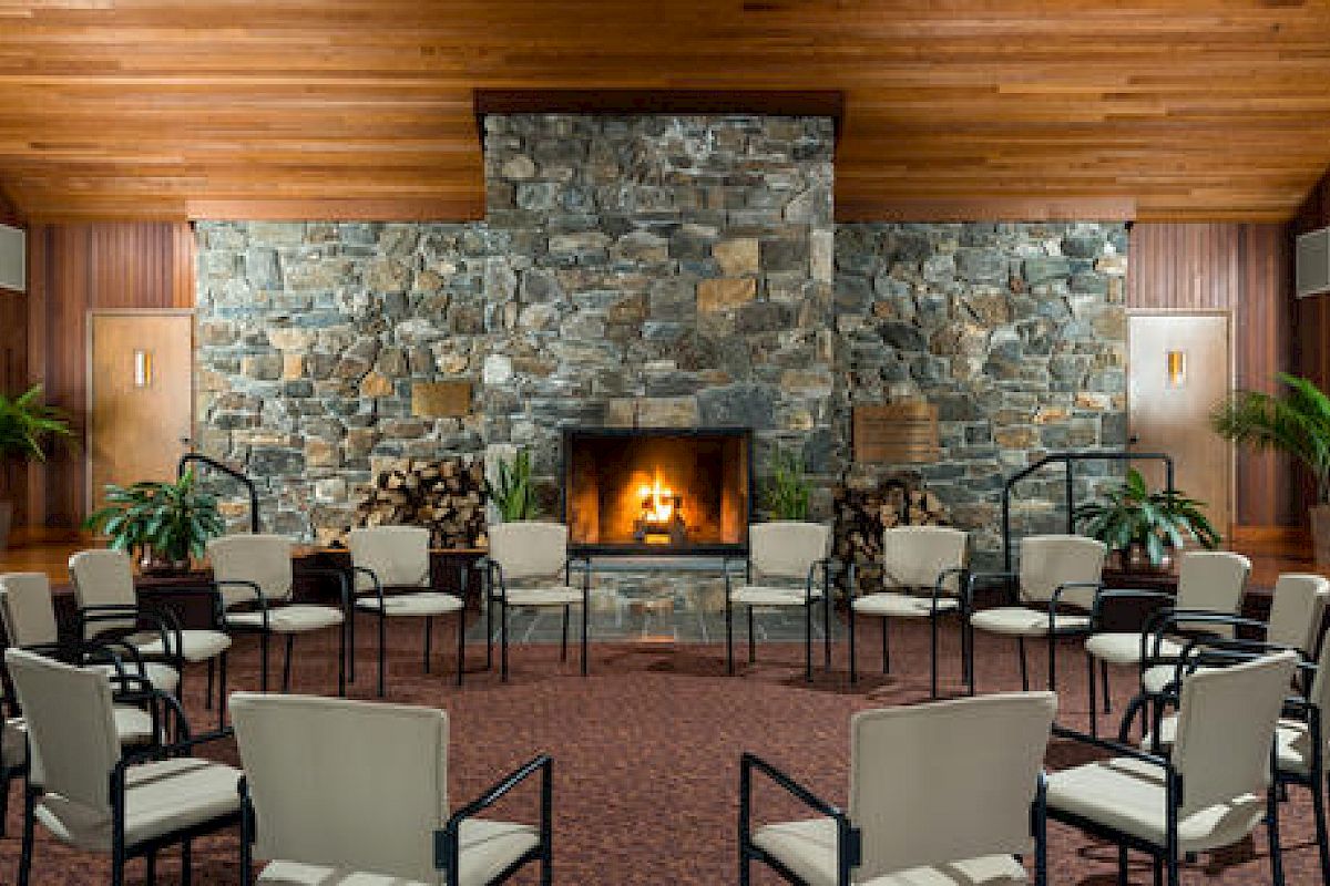 A cozy room with a lit fireplace, stone wall, wood ceiling, chairs arranged in a circle.