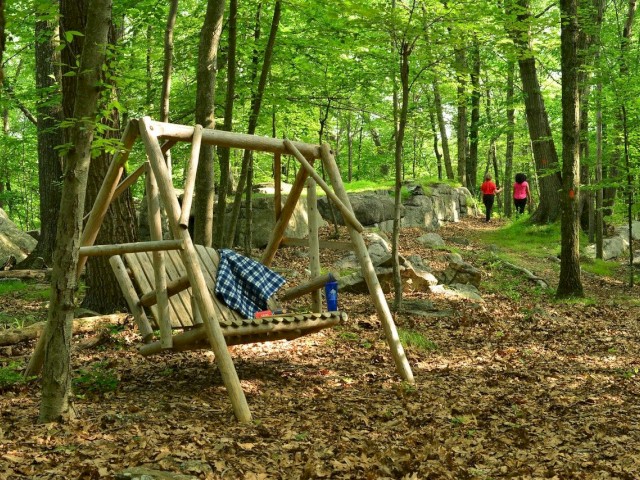 Wooden swing with a blanket in a forest; two people walking in the background.