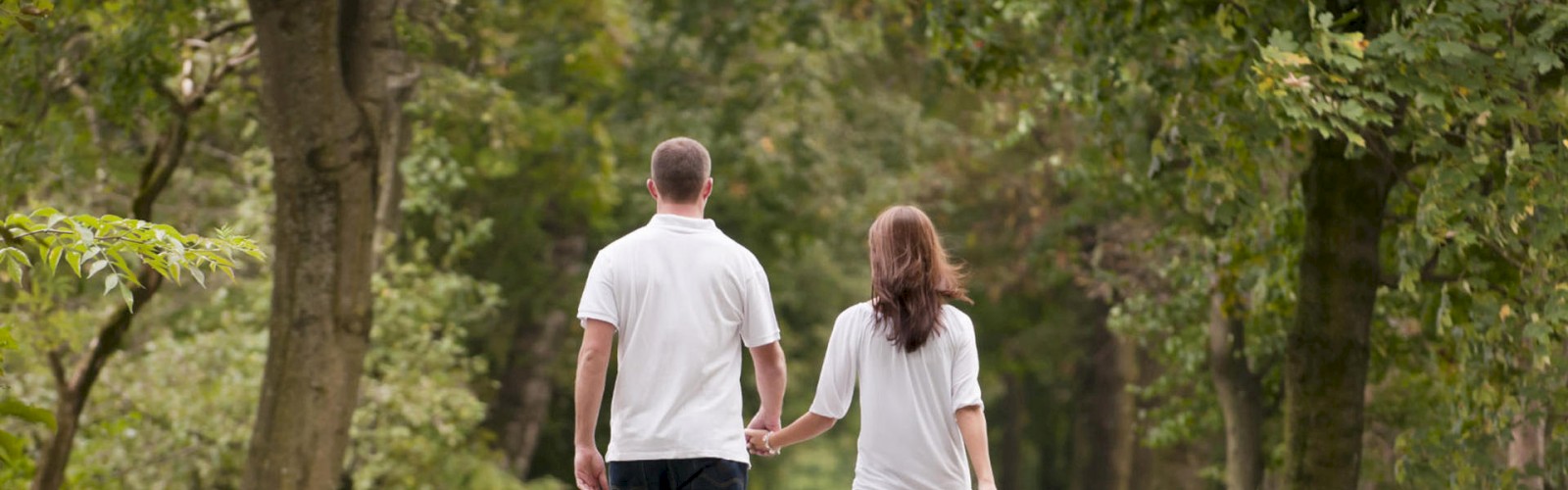 A couple is holding hands and walking on a tree-lined path in a park, both wearing white shirts, surrounded by greenery and natural scenery.