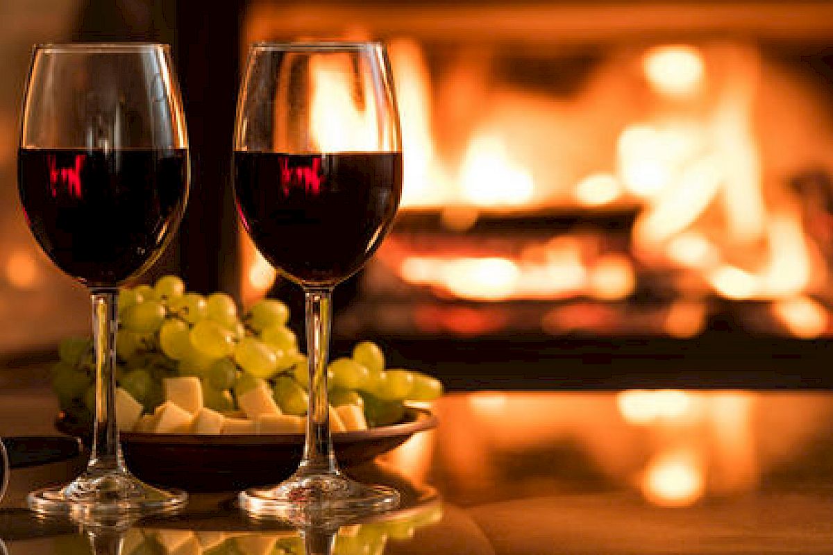 The image shows two glasses of red wine, a plate with green grapes and cheese, and a warm fireplace in the background.