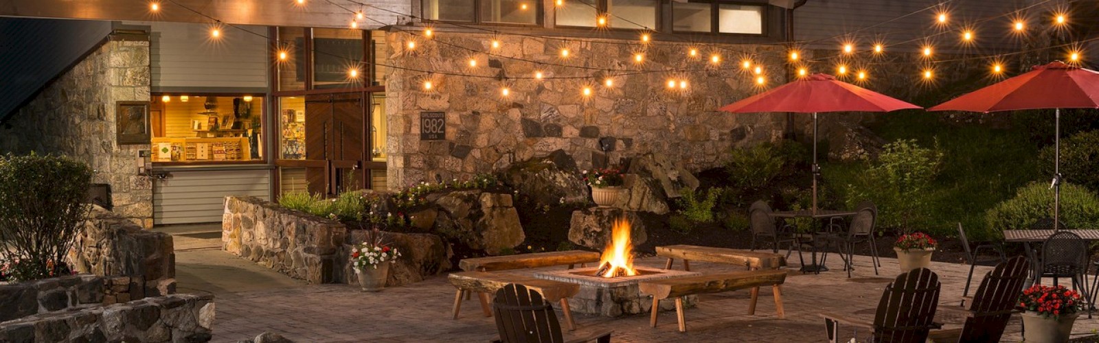Cozy backyard patio at dusk with fire pit, string lights, chairs, and a house with interior lights on.
