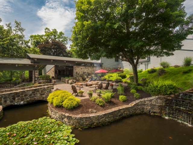 A serene outdoor space with a patio, tables, chairs, and a red umbrella, surrounded by lush greenery and a pond.