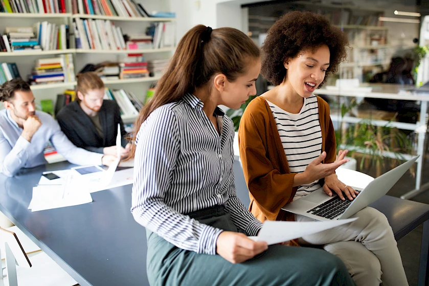 Two women are looking at a laptop and discussing, with others working in the background.