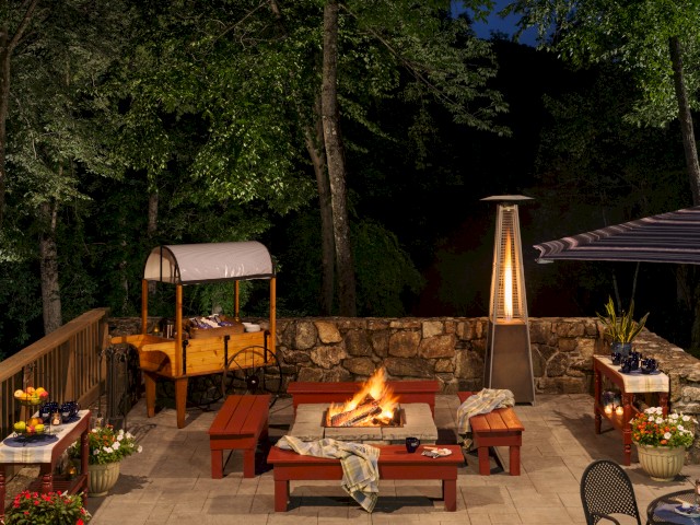 A cozy outdoor patio with a fire pit, benches, a tall heater, dining tables, and lush trees surrounding the area at night.
