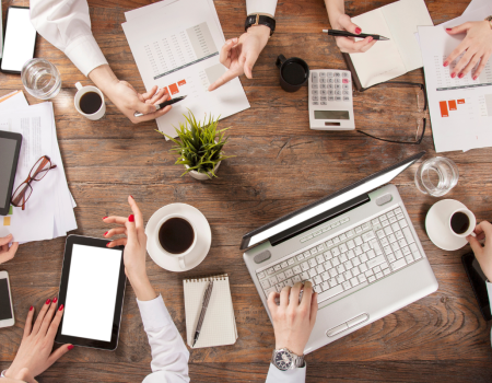 A group of people at a wooden table, working with laptops, tablets, documents, and a calculator; there's also coffee and a potted plant on the table.