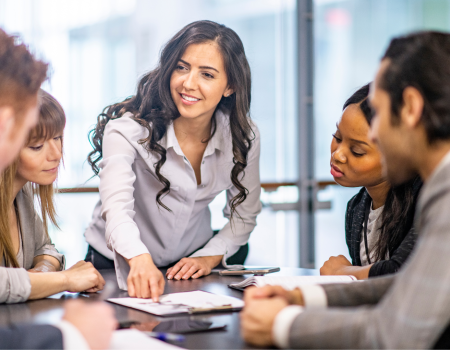 A group of five people are seated around a table, engaged in a discussion. One person is standing, pointing at a document on the table.