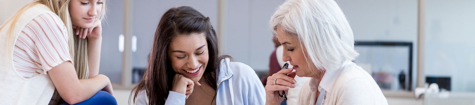 Three women are engaged in a discussion at a table with a laptop and documents. They're in a well-lit, modern classroom or office space.