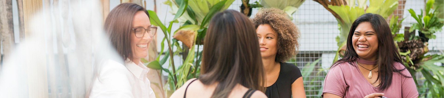 Four women are sitting at a table outdoors, engaging in a lively conversation, surrounded by lush greenery and plants.