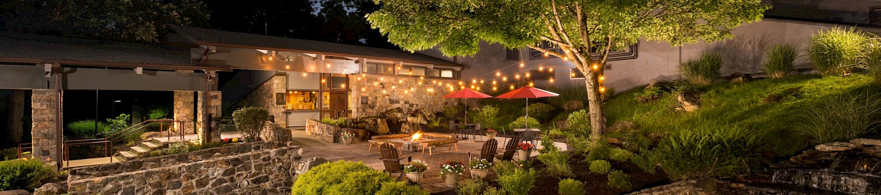 A serene outdoor patio with string lights, lush greenery, a pond, and comfortable seating under a night sky.