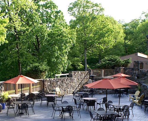 The image shows an outdoor patio with tables, chairs, and orange umbrellas, surrounded by lush green trees and stone walls.
