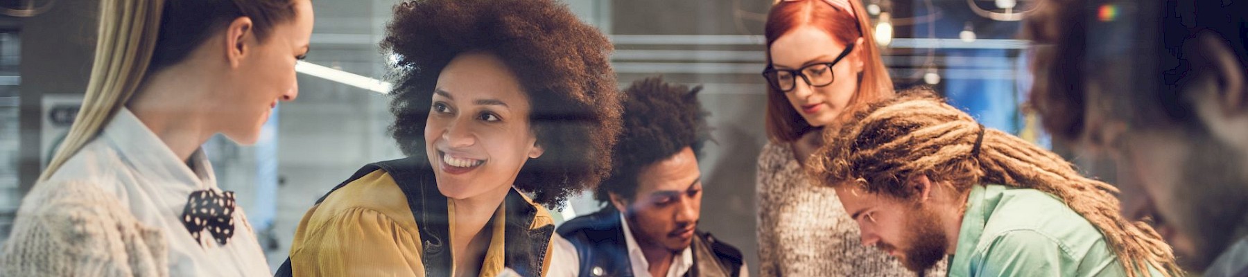 A group of people are gathered around a table discussing documents and using digital devices in a collaborative setting.