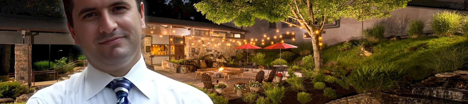 A man in a white shirt and tie stands confidently; behind him is a beautifully lit outdoor patio area during the evening, surrounded by trees.