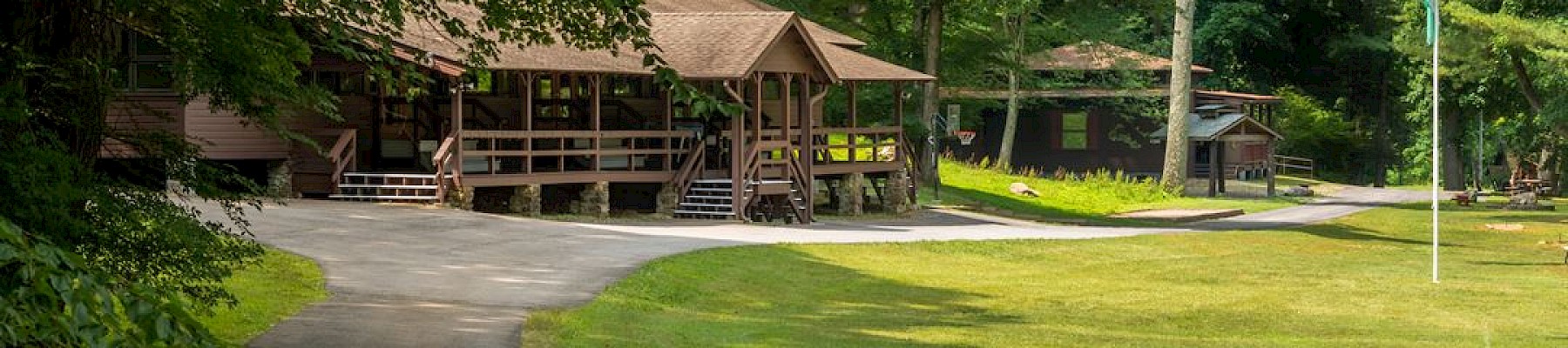 A peaceful outdoor scene with a pathway leading to a wooden cabin surrounded by trees and a grassy area with a flagpole.