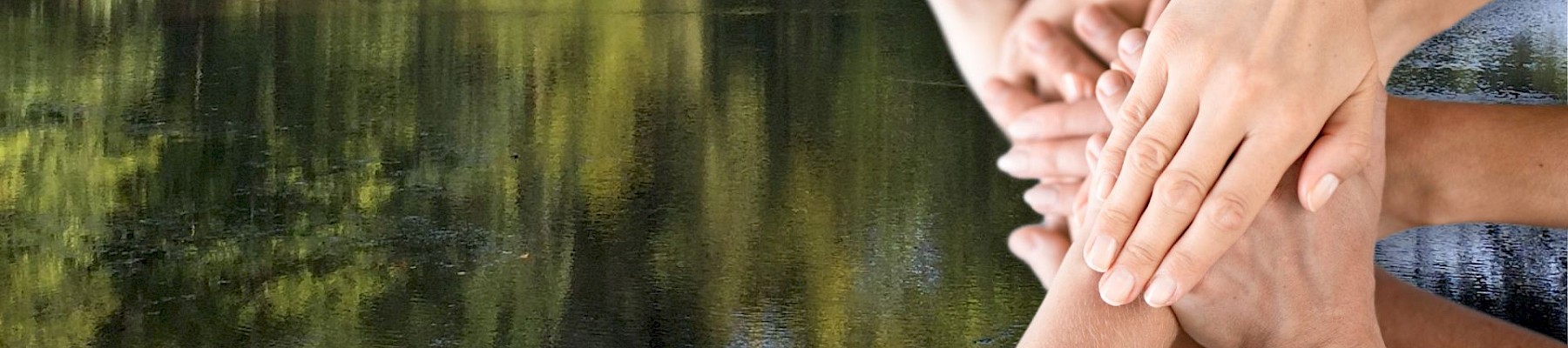 Several hands stacked together in a gesture of unity, set against a backdrop of a serene lake and lush trees.