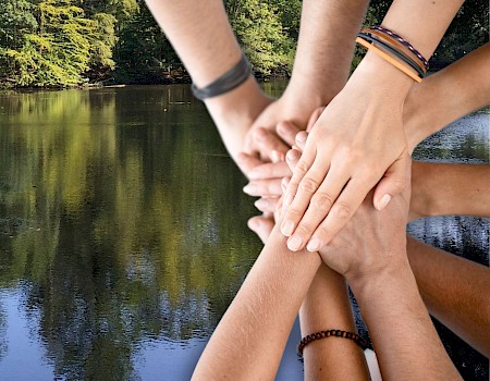 Several hands stacked together in a gesture of unity, set against a backdrop of a serene lake and lush trees.