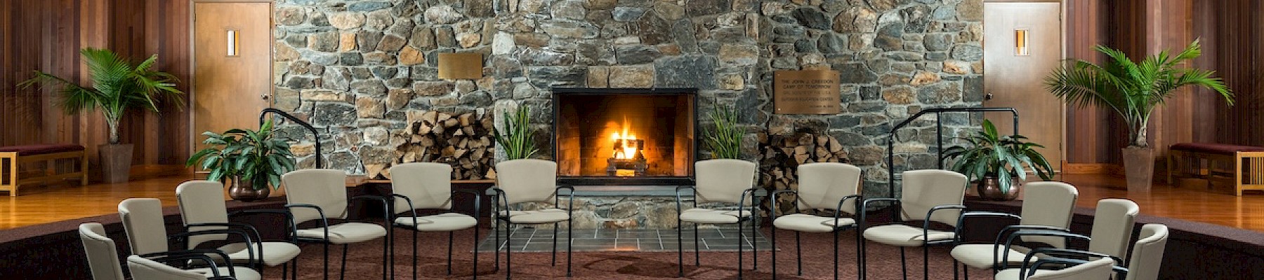 A room with a circle of chairs, a stone fireplace, wooden ceiling, and potted plants, creating a cozy and inviting atmosphere.