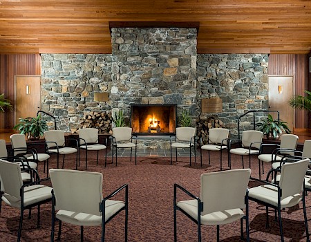 A room with a circle of chairs, a stone fireplace, wooden ceiling, and potted plants, creating a cozy and inviting atmosphere.