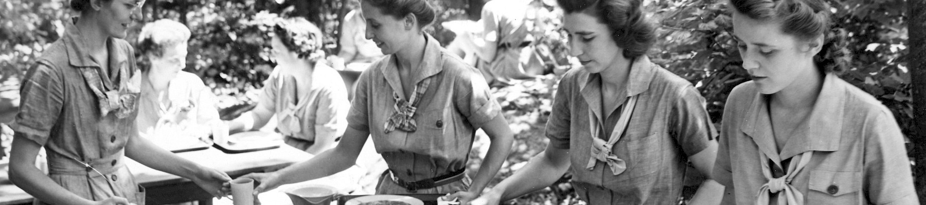 A group of women in uniforms is serving food buffet-style outdoors, with various dishes arranged on a table in a wooded setting.