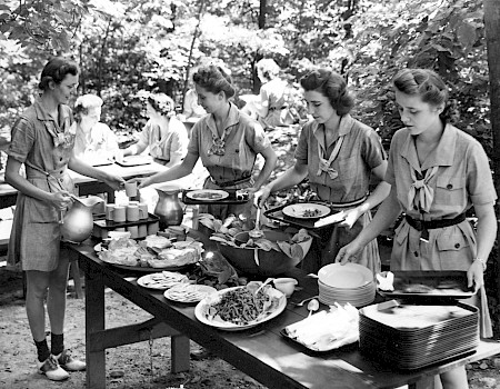 A group of women in uniforms is serving food buffet-style outdoors, with various dishes arranged on a table in a wooded setting.