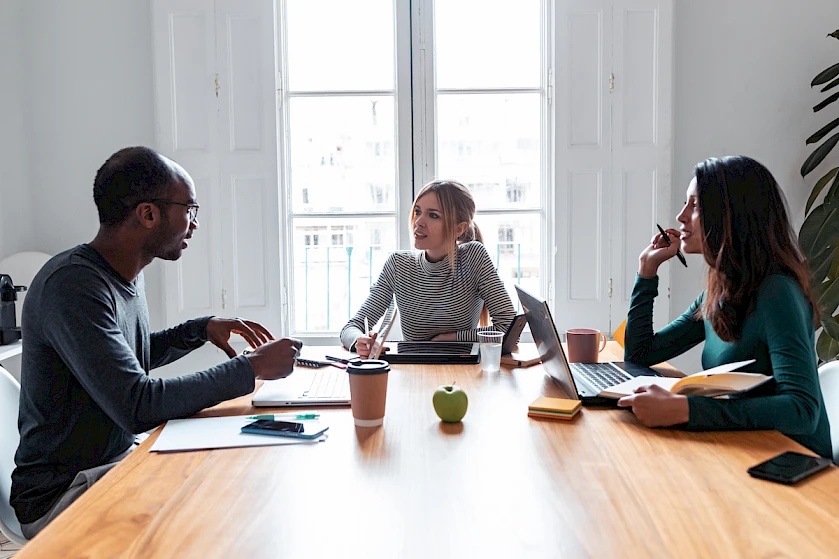 Three people are in a meeting at a table with laptops and an apple.